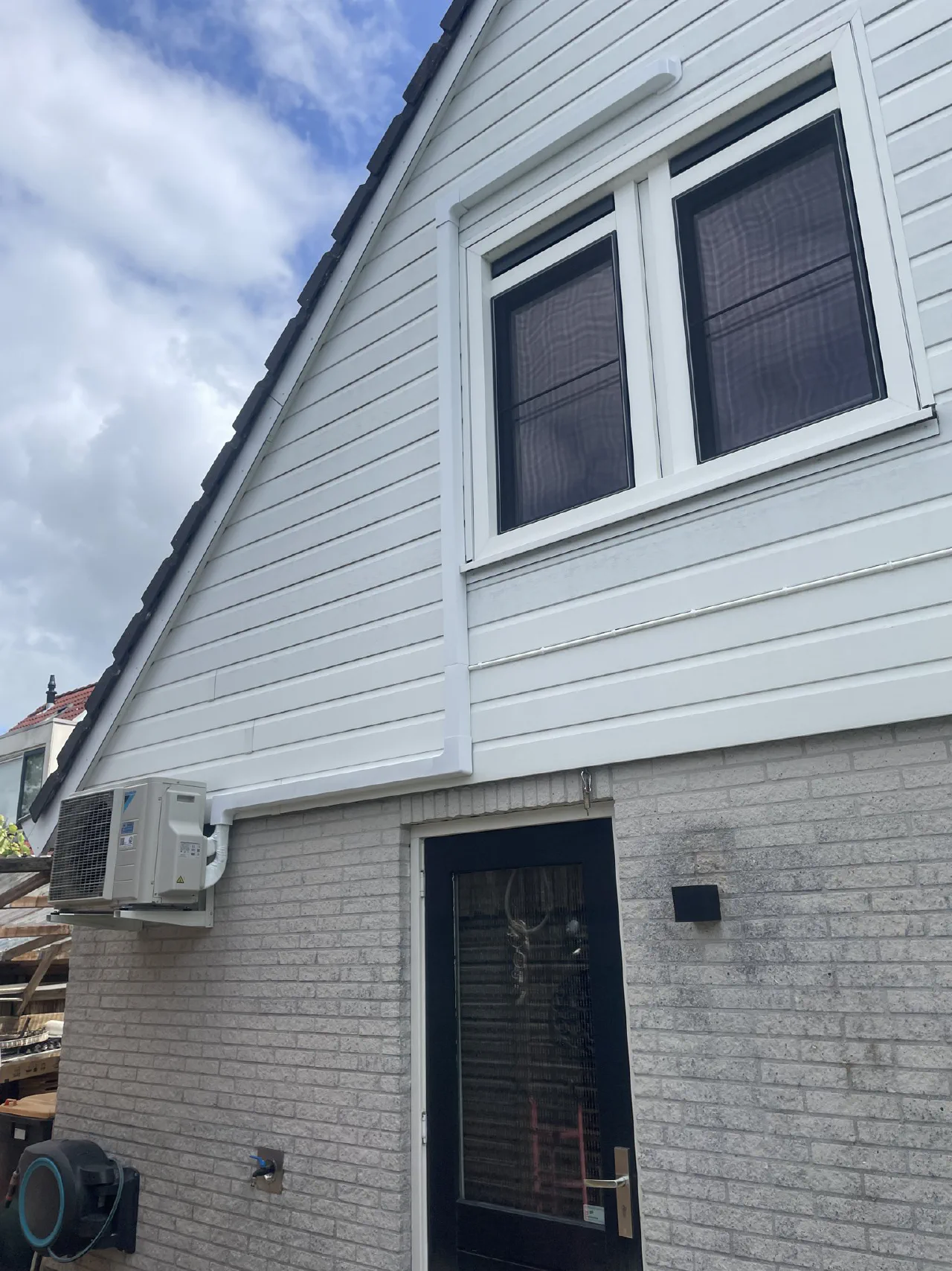 Witte buitenunit op een muurbeugel aan een lichte bakstenen achtergevel naast een zwarte achterdeur, onder een witte houten puntgevel met raam, witte leidinggoot langs de gevelbekleding
