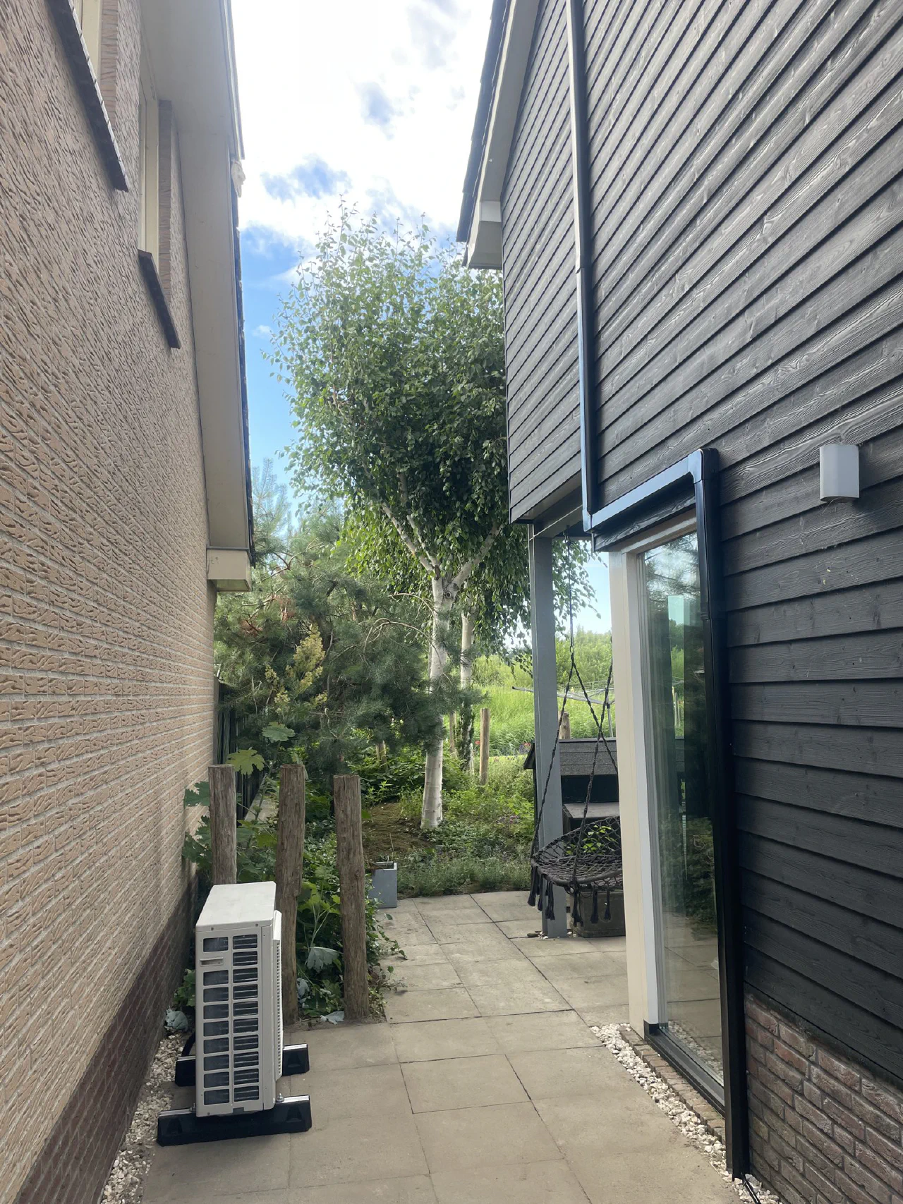Witte buitenunit in een smalle betegelde doorgang tussen een lichte bakstenen gevel en een woning met zwarte houten gevelbekleding, zwarte leidinggoot langs het raam omhoog