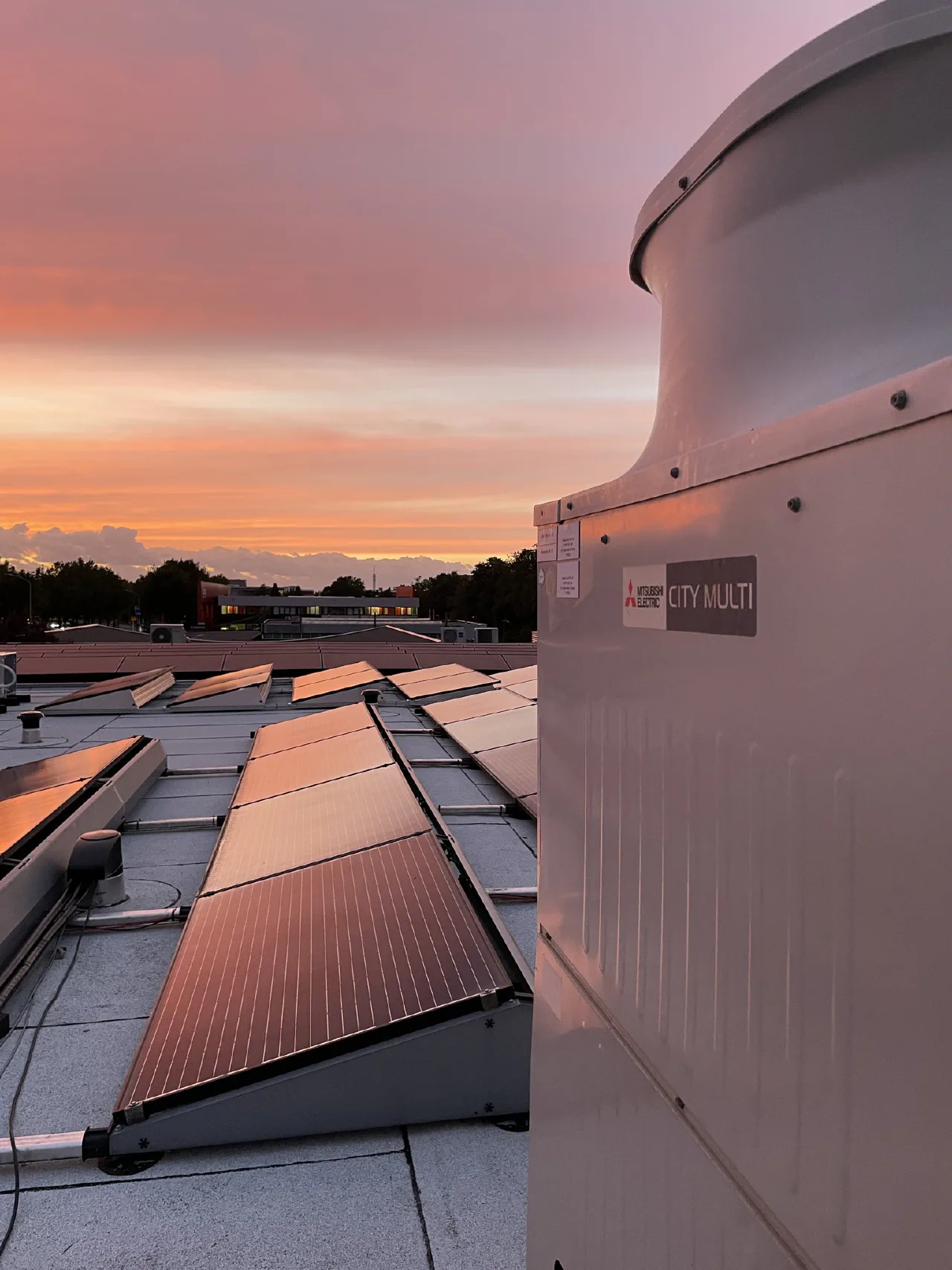Close-up van het City Multi logo op de buitenunit, met zonnepanelen en een roze avondlucht op de achtergrond
