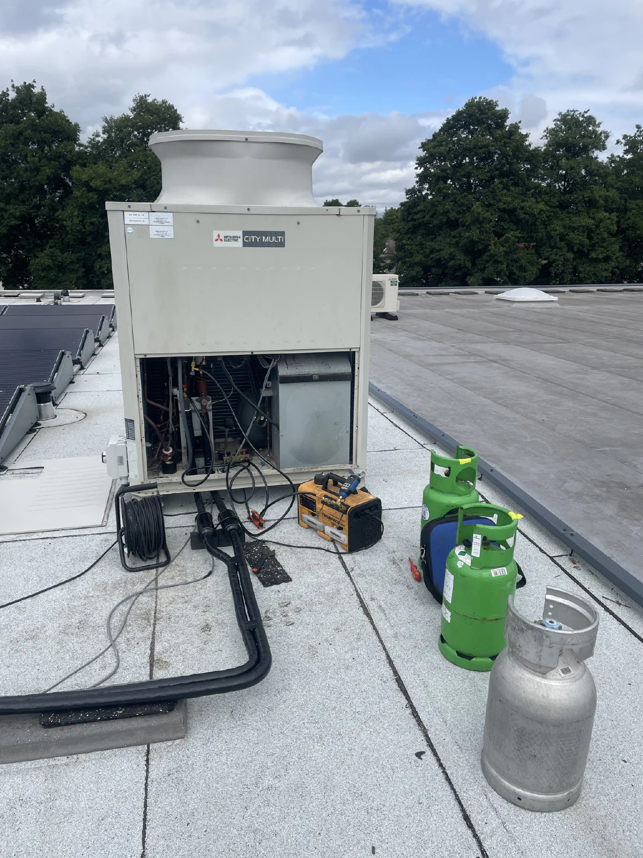 Geopende Mitsubishi Electric City Multi buitenunit op een plat dak met zonnepanelen, ervoor een vacuümpomp, een haspel en groene koudemiddelcilinders
