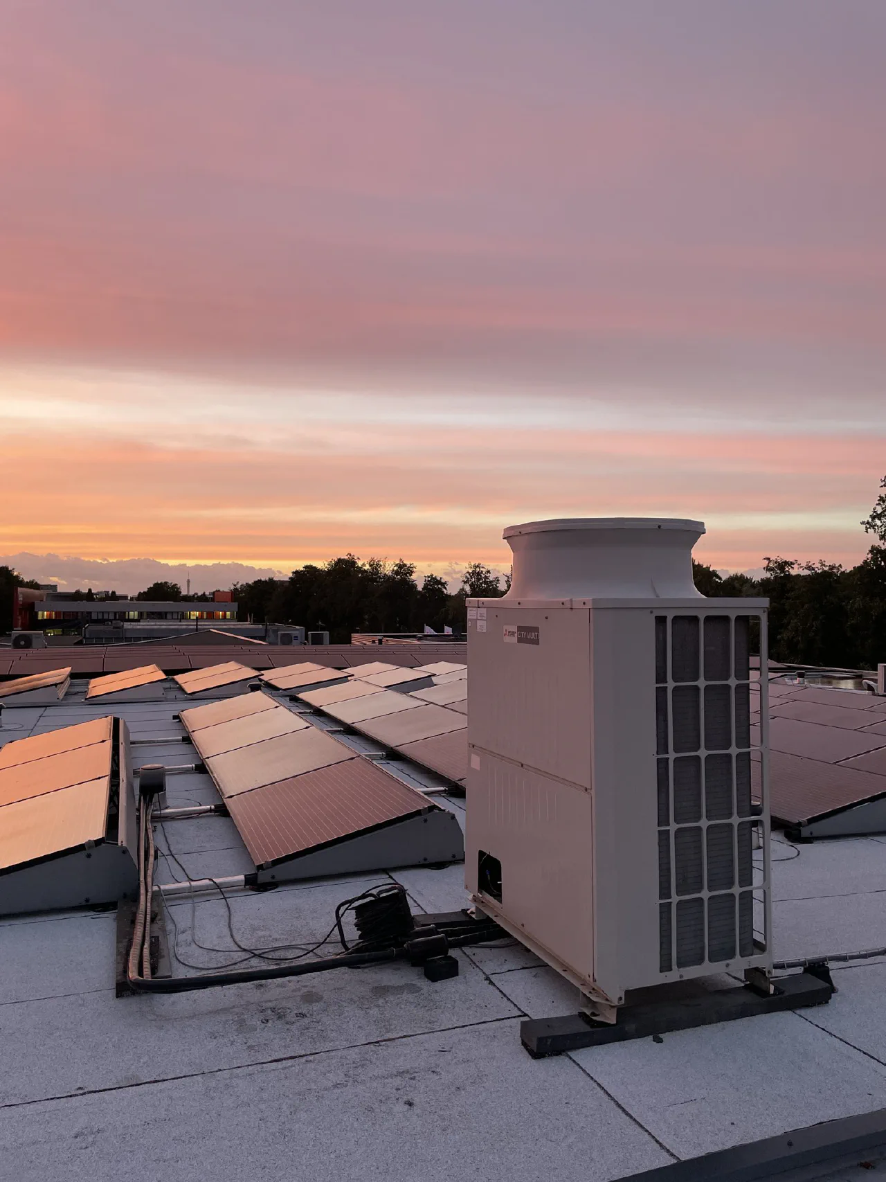 Mitsubishi Electric City Multi buitenunit op een plat dak met rijen zonnepanelen bij een roze-oranje zonsondergang