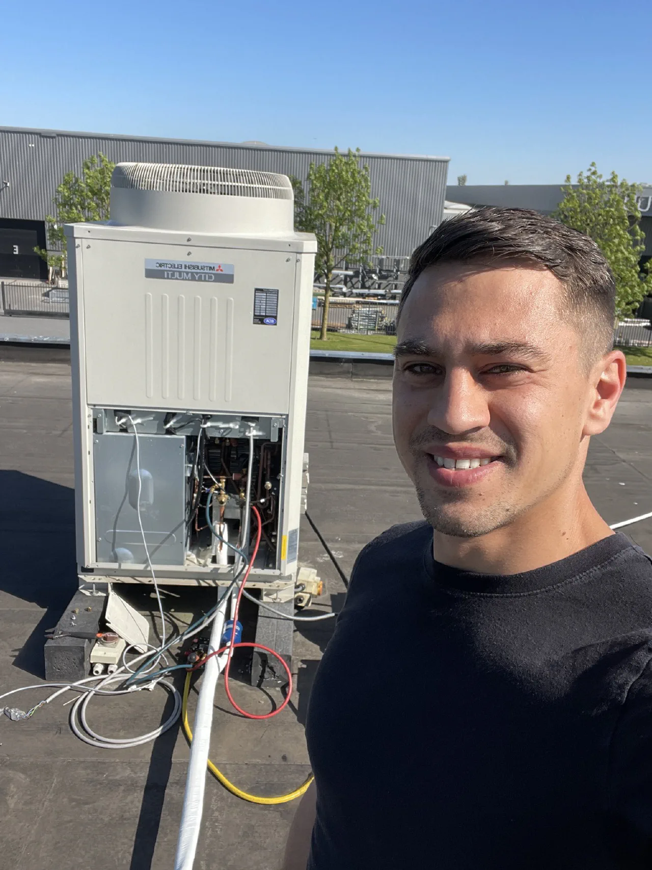 Selfie van de installateur op een plat dak naast een geopende Mitsubishi Electric City Multi buitenunit met aangesloten leidingen en kabels, bedrijfshal op de achtergrond