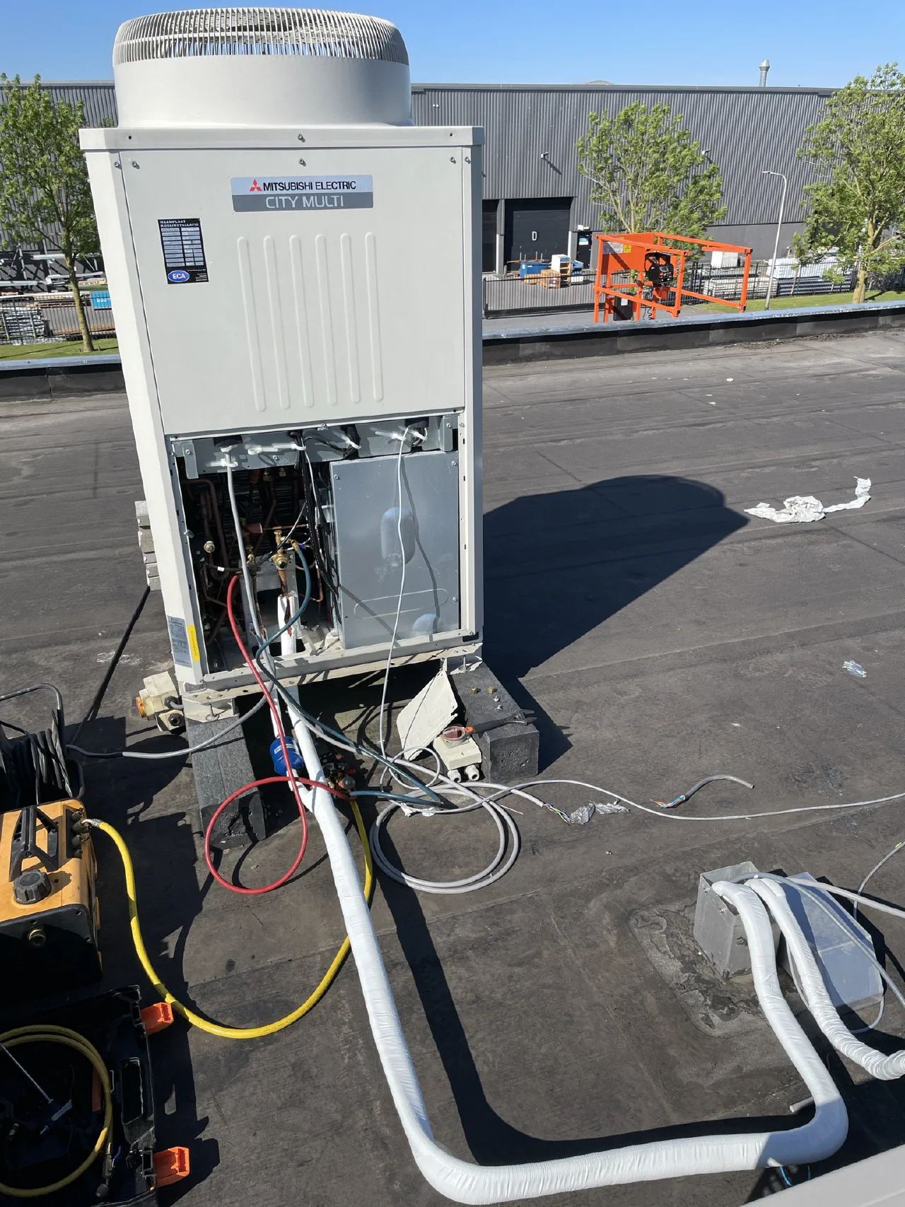 Geopende Mitsubishi Electric City Multi buitenunit op het dak van een bedrijfspand, met aangesloten geïsoleerde leidingen, bekabeling en een vacuümpomp met gele slang, oranje hoogwerker op de achtergrond