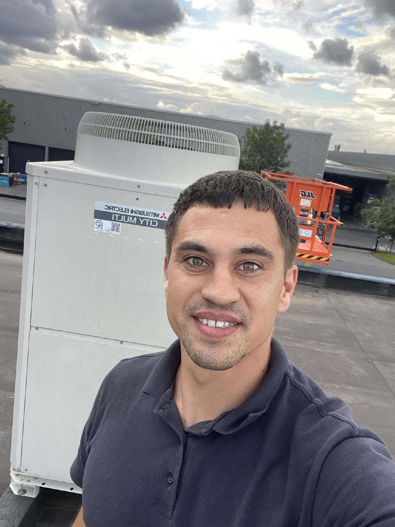 Selfie van de installateur op het dak voor de Mitsubishi Electric City Multi buitenunit, met een oranje hoogwerker en bedrijfshallen op de achtergrond