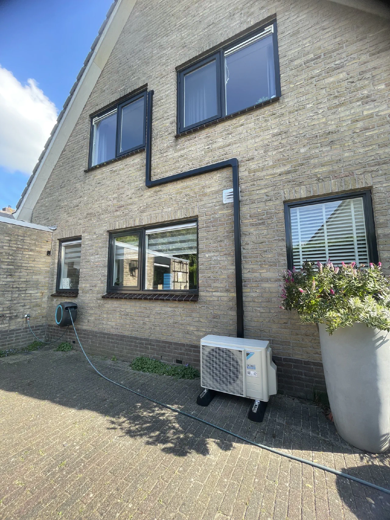 Daikin buitenunit op een oprit tegen een gele bakstenen gevel, antraciete leidinggoot loopt in trapvorm tussen de ramen door naar de verdieping