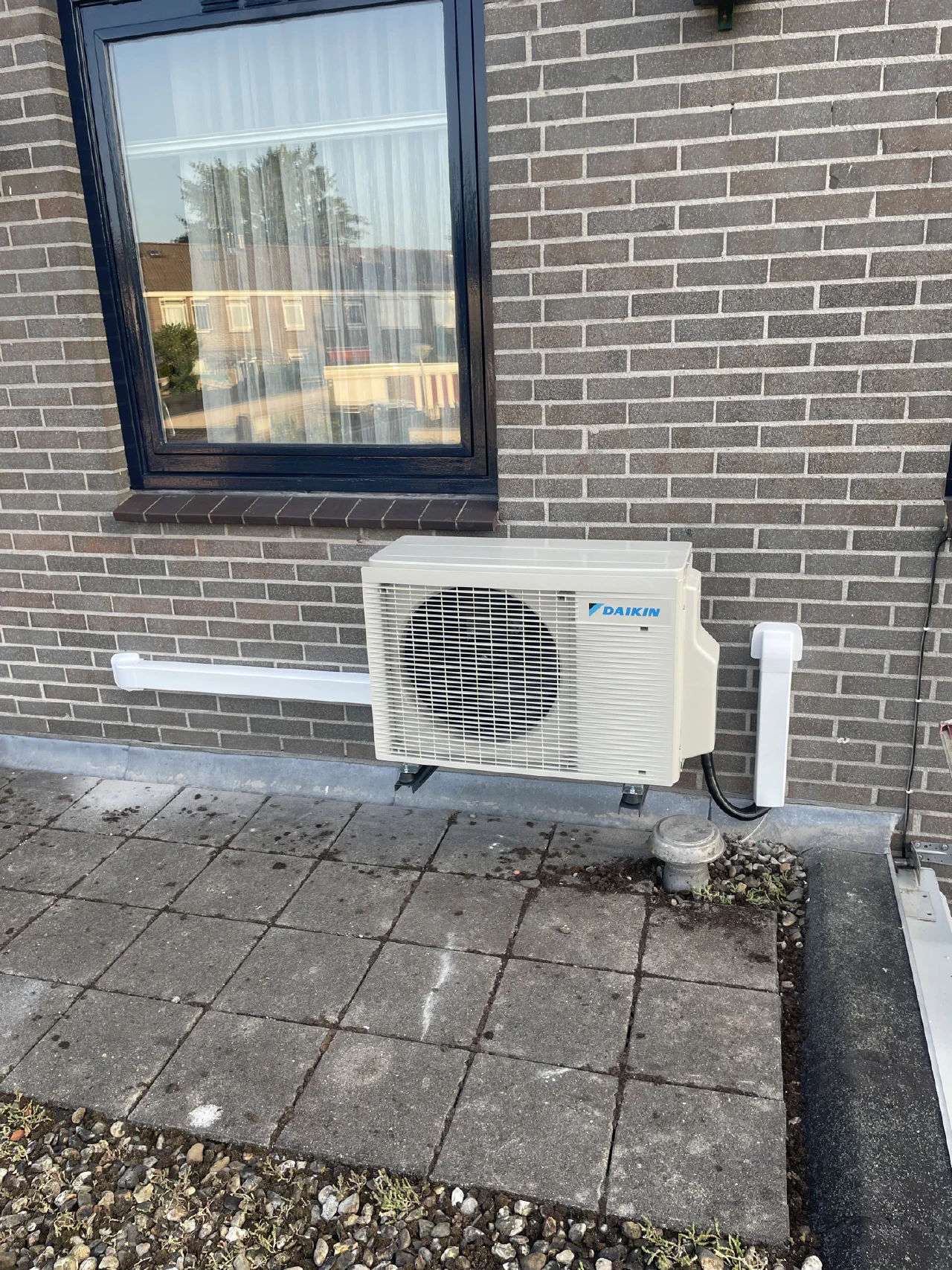 Daikin buitenunit op rubber voeten op een plat dak met betontegels, tegen een grijze bakstenen gevel onder een raam, witte leidinggoten links en rechts