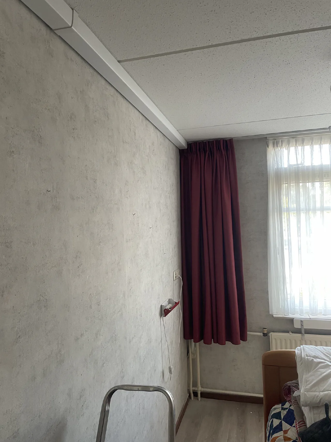 Slaapkamer met grijze wand, witte leidinggoot langs het plafond, bordeauxrode gordijnen, een raam en een aansluitpunt op de wand