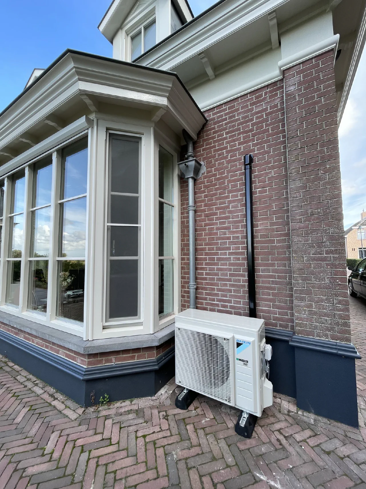 Daikin buitenunit op dempers naast de erker van een klassieke woning met rode baksteen, crèmekleurig houtwerk en donkere plint, zwarte leidinggoot omhoog naast een regenpijp