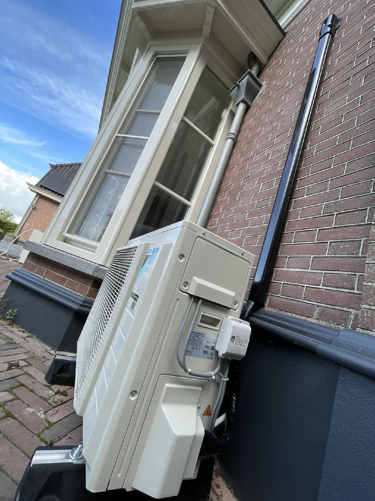 Close-up van de Daikin buitenunit van opzij, met aansluitkast, zwarte leidinggoot omhoog langs de gevel en het raam van de erker erboven