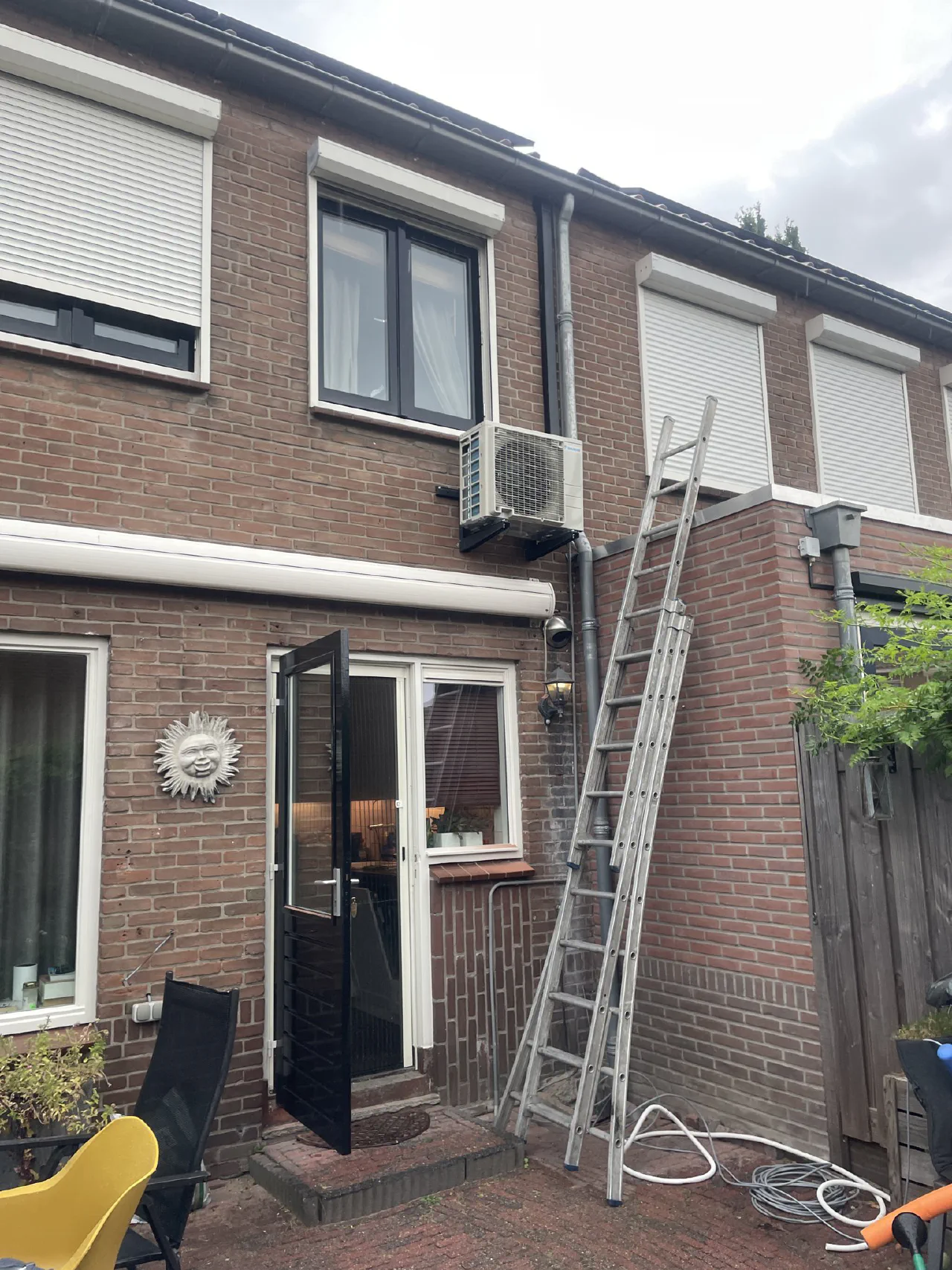 Daikin buitenunit op een zwarte muurbeugel aan een bakstenen achtergevel boven een zonnescherm, met een ladder tegen de gevel en losse leidingen op de grond