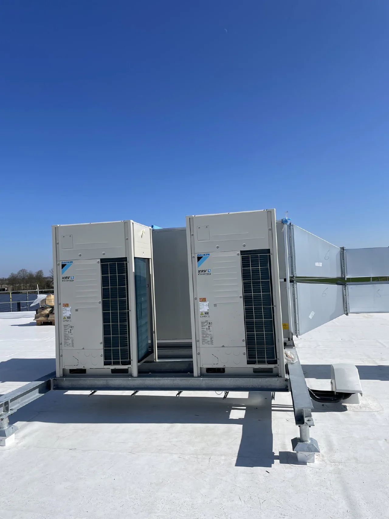 Twee Daikin VRV 5 inverter buitenunits op een gegalvaniseerd stalen frame op een wit plat dak, met rechts een luchtbehandelingskast met kanaalwerk, strakblauwe lucht