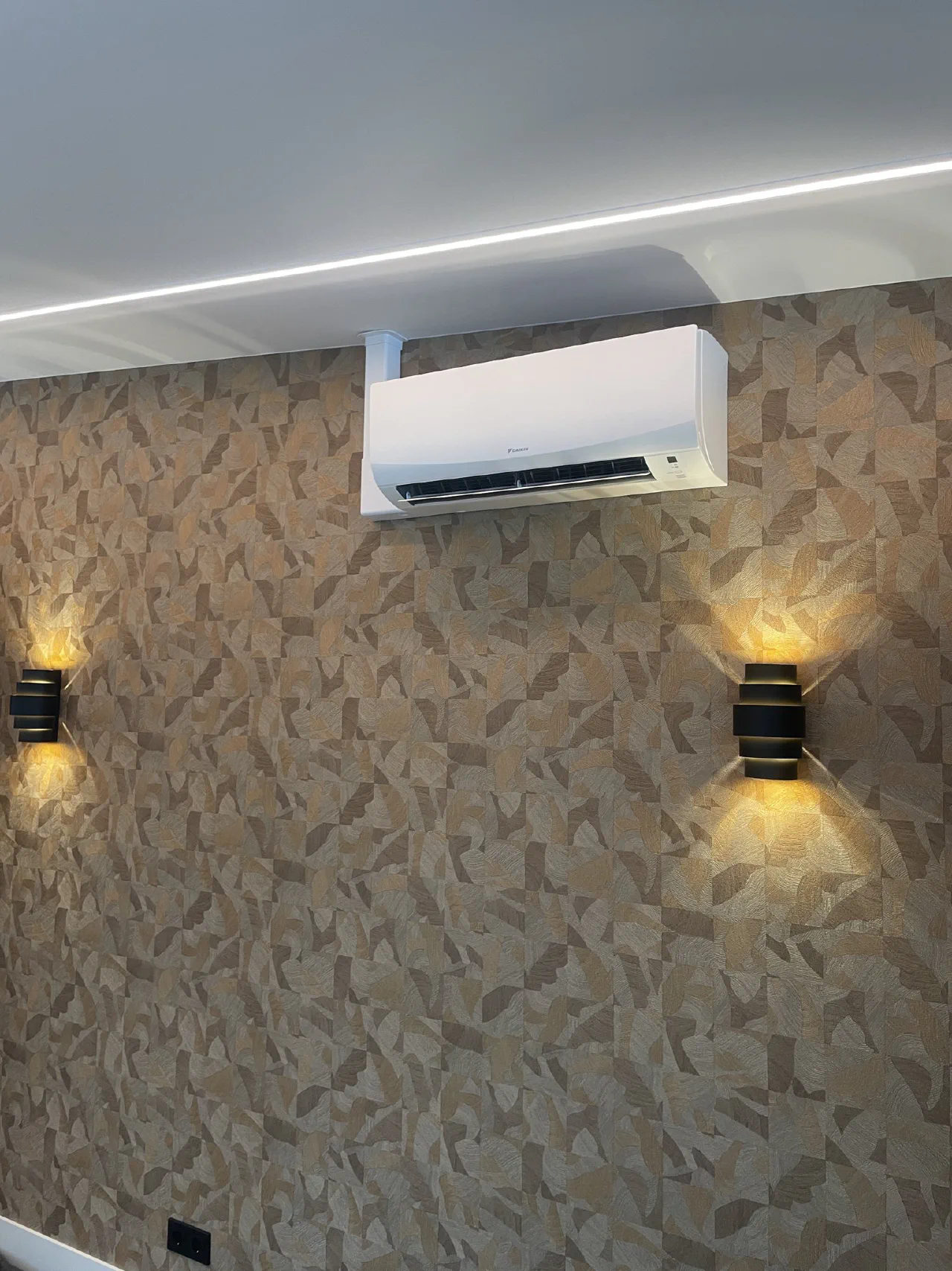 Witte Daikin wandunit op een behangen wand tussen twee zwarte wandlampen, met een LED-lichtlijn in het plafond
