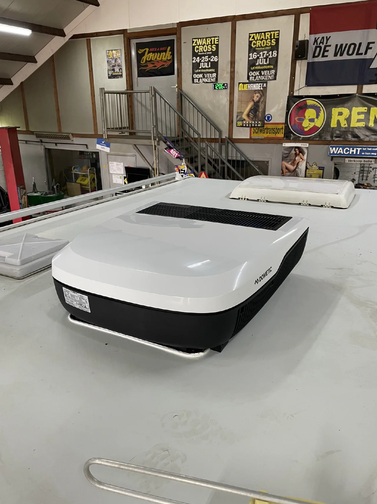 Witte Dometic dakairco op het vlakke witte dak van een camper in een werkplaats, met een dakluik erachter en posters aan de wand