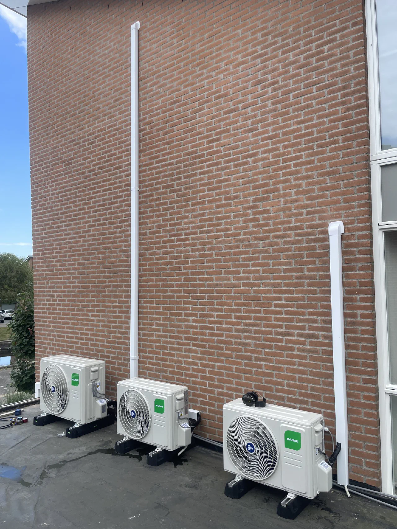 Drie witte Kaisai buitenunits naast elkaar op een dakterras tegen een hoge rode bakstenen gevel, met twee witte leidinggoten omhoog