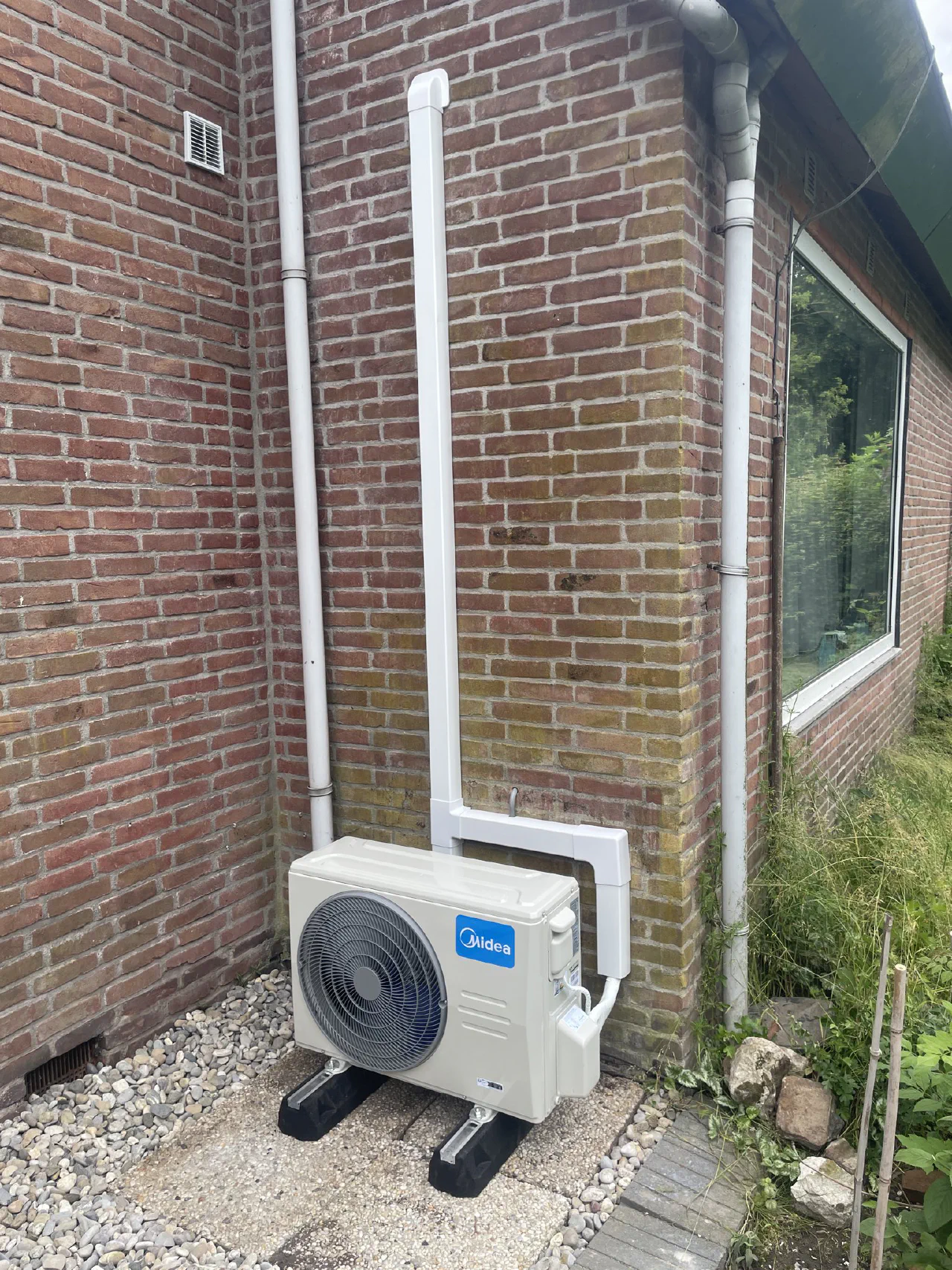 Midea buitenunit op grind in de hoek van een rode bakstenen woning, witte leidinggoot omhoog naast een witte regenpijp