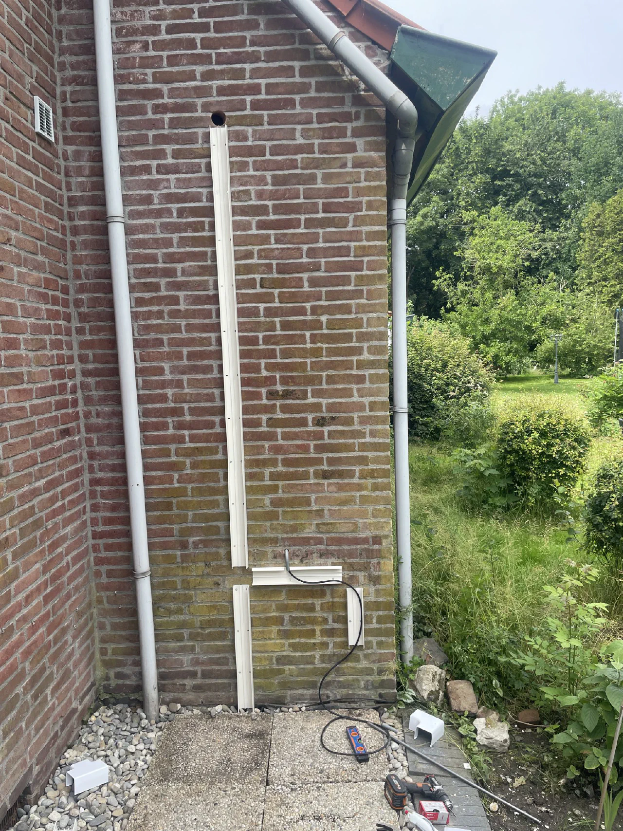 Achterstuk van een witte leidinggoot recht onder een geveldoorvoer in rode baksteen, tussen een ventilatierooster en een regenpijp, gereedschap op de tegels