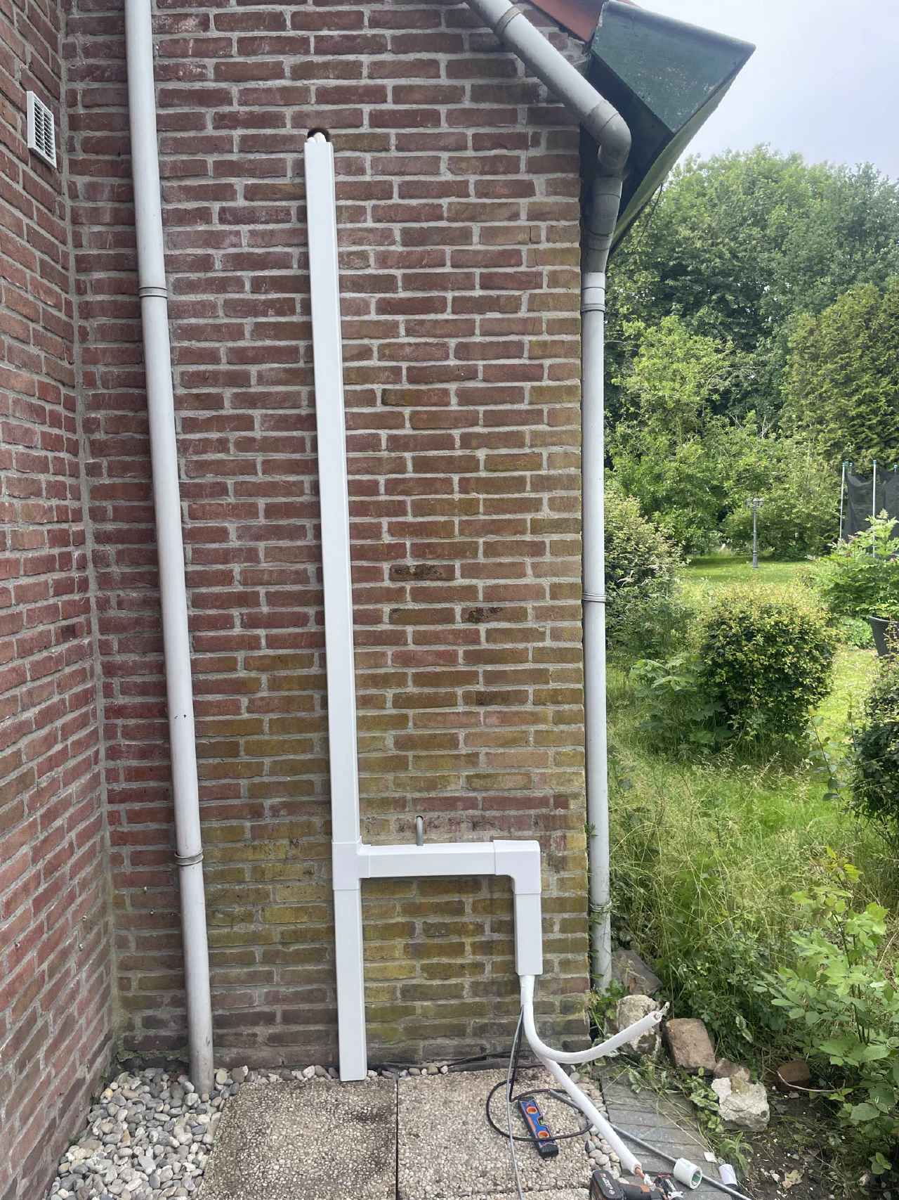 Witte leidinggoot met haakse bocht langs een rode bakstenen gevel naast een regenpijp, kabels komen onderaan uit de goot