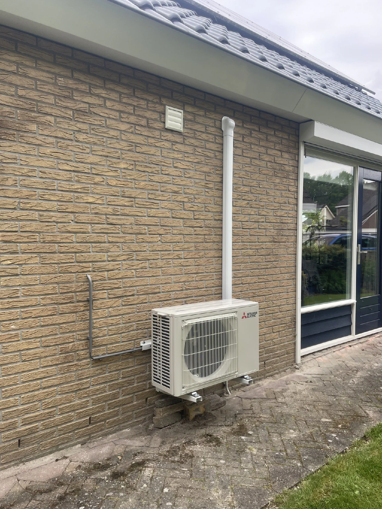 Mitsubishi Electric buitenunit op rubber voeten tegen een gele bakstenen gevel, witte leidinggoot omhoog naar de dakrand, grijze voedingsbuis links