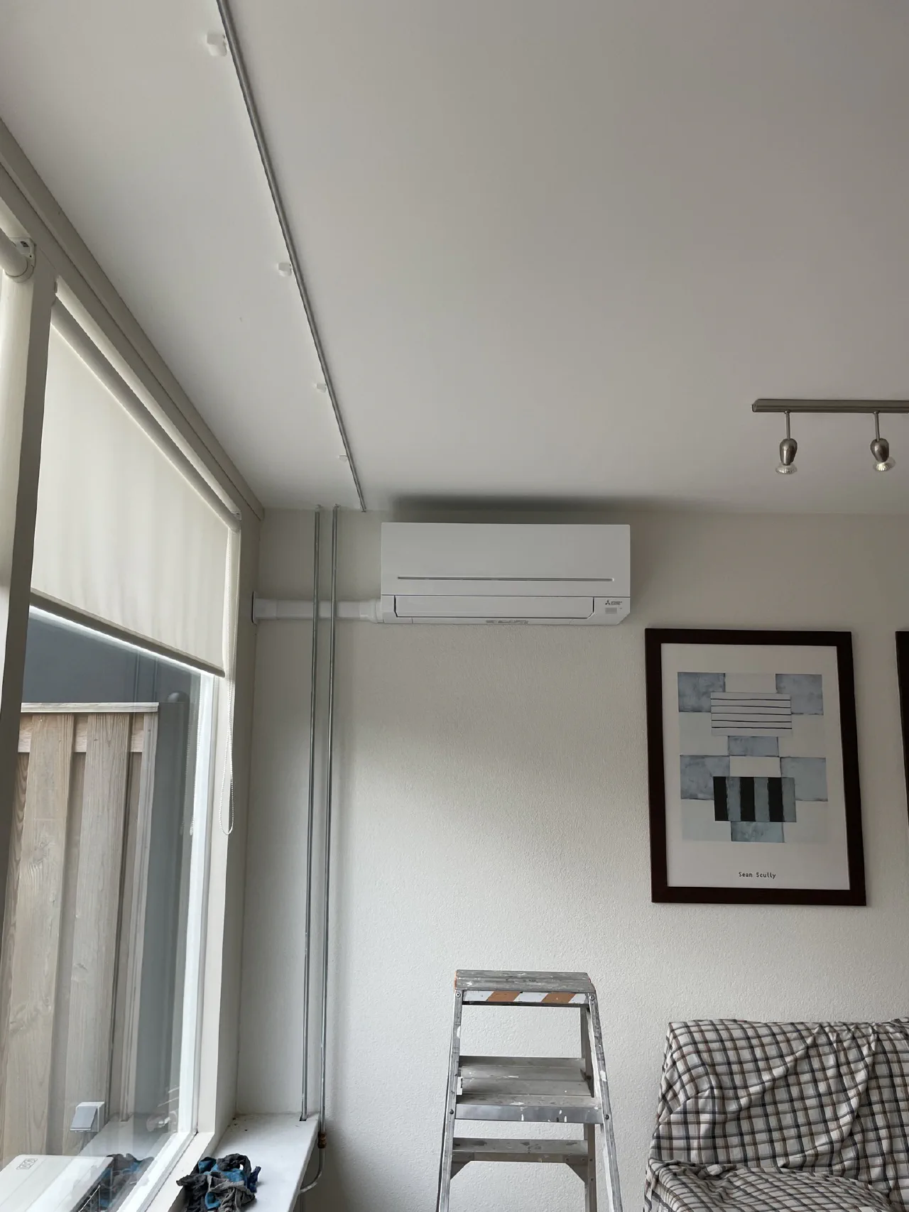 Witte Mitsubishi Electric wandunit hoog in de hoek van een woonkamer naast het raam, grijze leidingbuizen langs het kozijn, een trap en afgedekte bank in de kamer