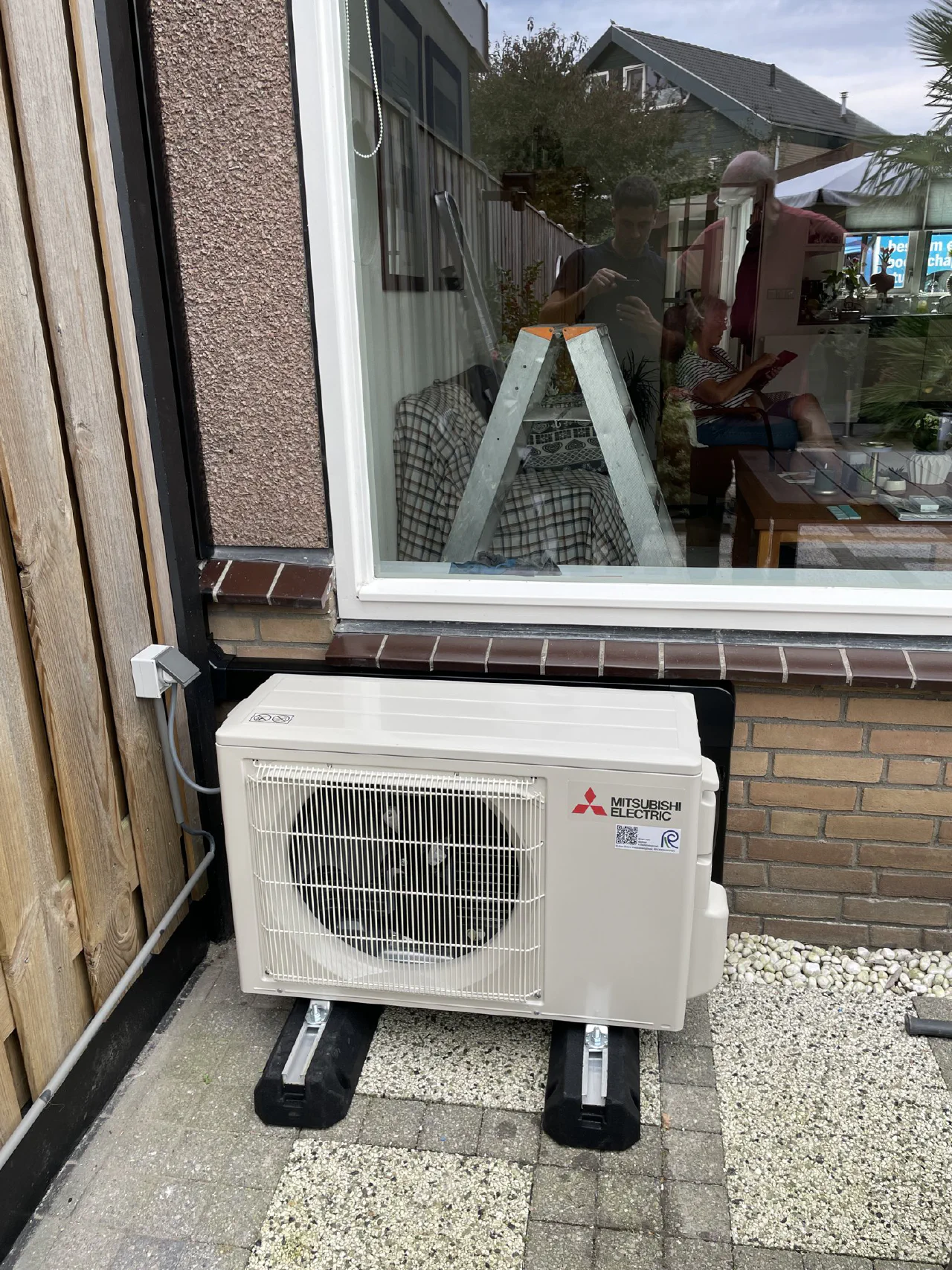 Mitsubishi Electric buitenunit op rubber voeten onder een groot raam, tegen een gevel van baksteen, stucwerk en houten schutting