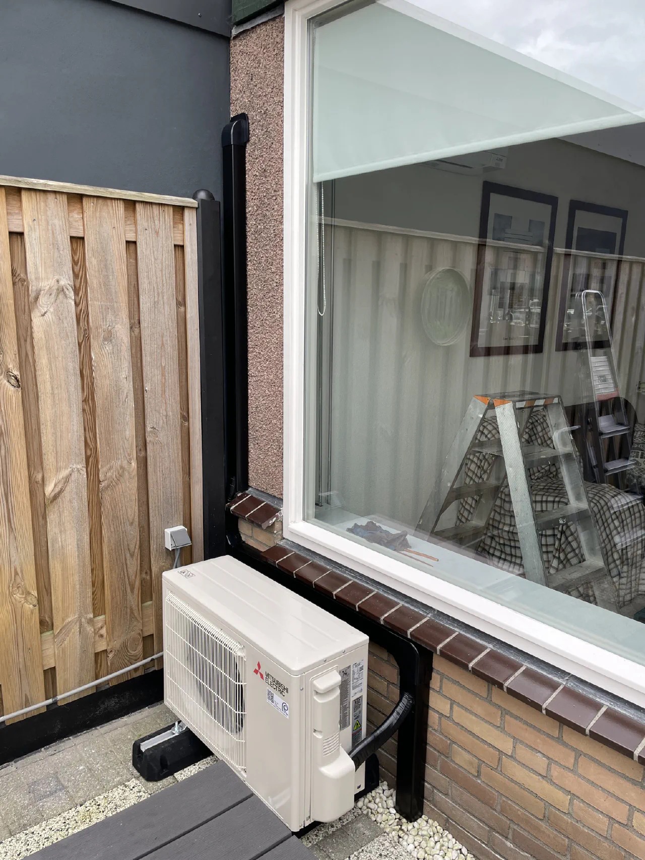 Mitsubishi Electric buitenunit naast een houten schutting, zwarte leidinggoot langs grijs stucwerk omhoog, grijze voedingsbuis links