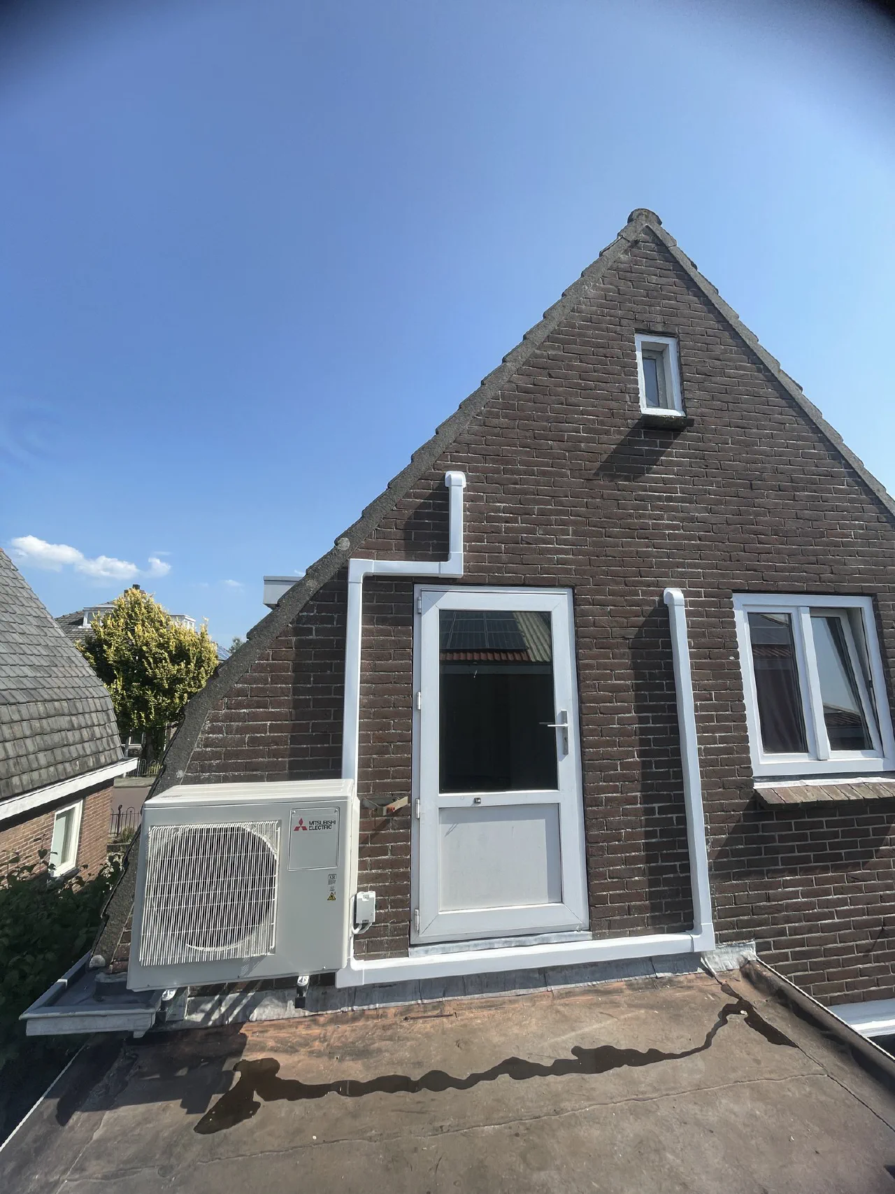 Mitsubishi Electric buitenunit op een beugel aan een donkere bakstenen puntgevel naast een witte deur, witte leidinggoten rond de deur, strakblauwe lucht