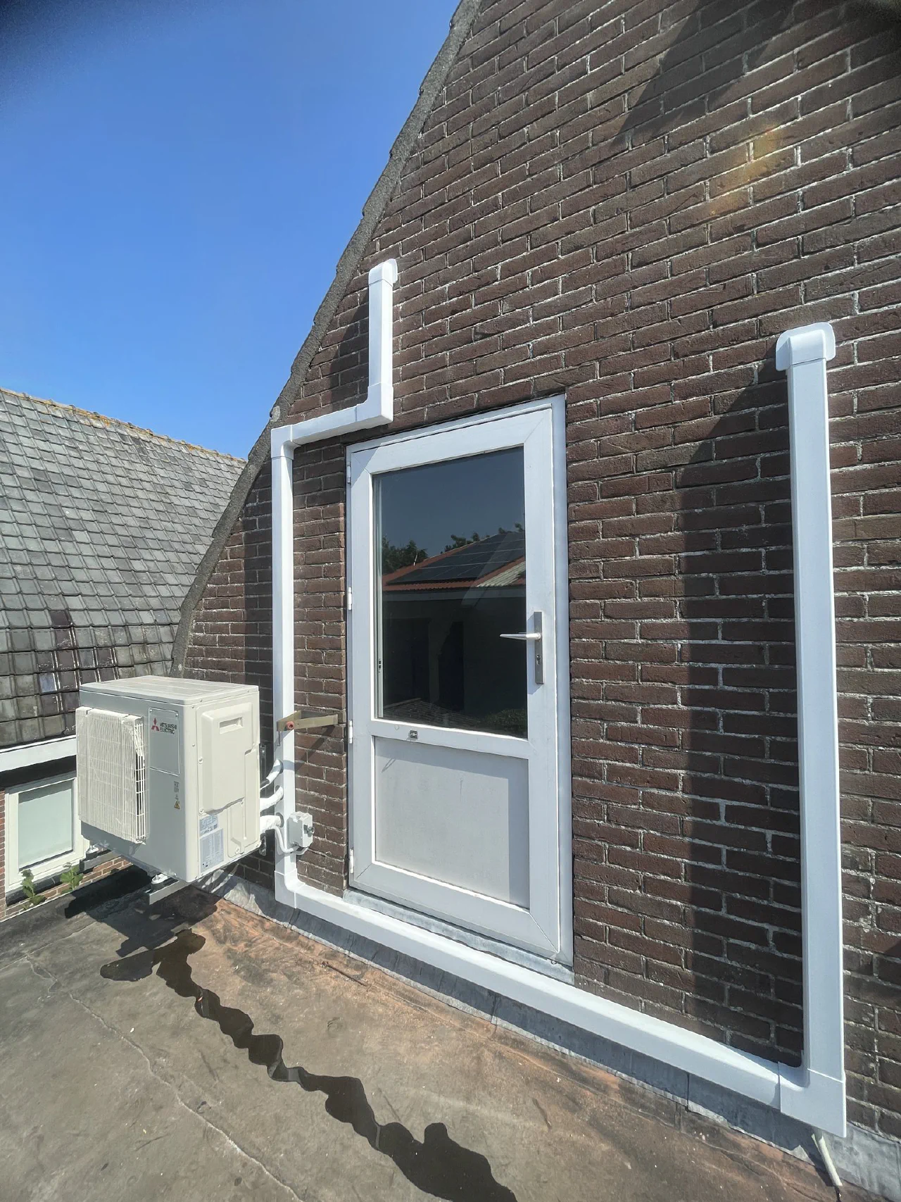 Zijaanzicht van de Mitsubishi Electric buitenunit aan de puntgevel, met witte leidinggoot boven de deur en langs de gevel omlaag