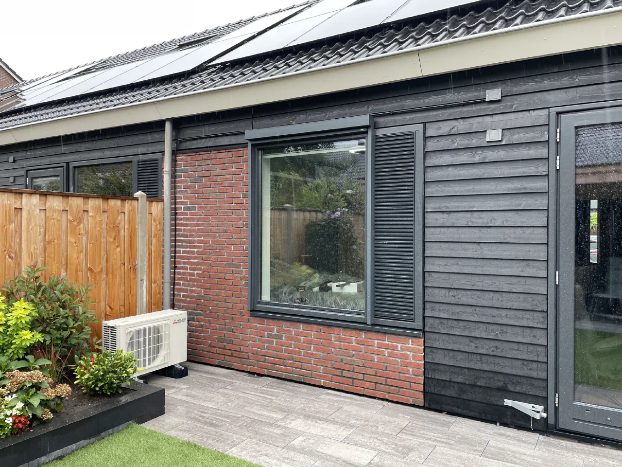 Mitsubishi Electric buitenunit op terrastegels naast een plantvak bij een bungalow met zwarte houten gevel, rode baksteen en zonnepanelen op het dak