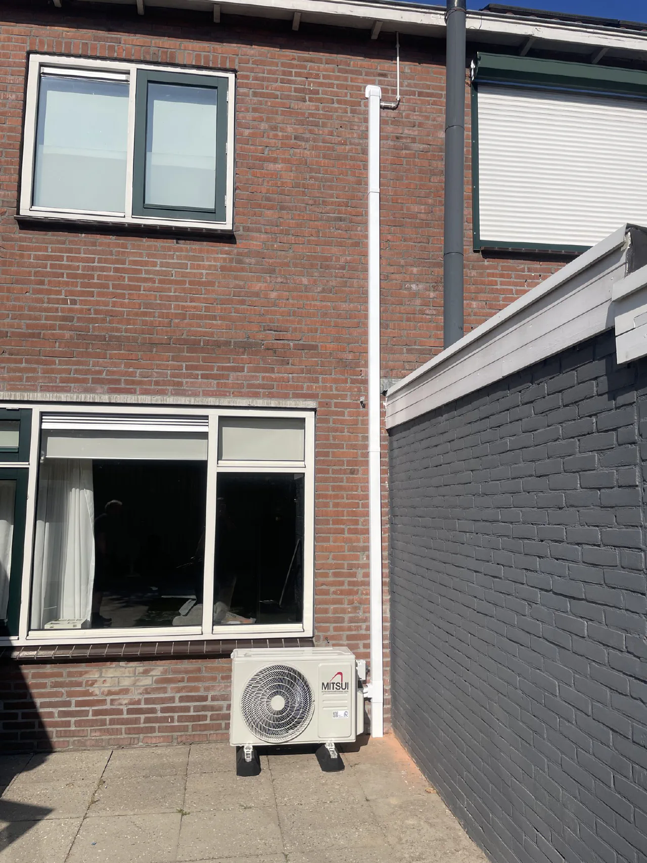 Mitsui buitenunit op de achterplaats van een rijtjeswoning tegen een rode bakstenen gevel, witte leidinggoot recht omhoog naar de dakrand