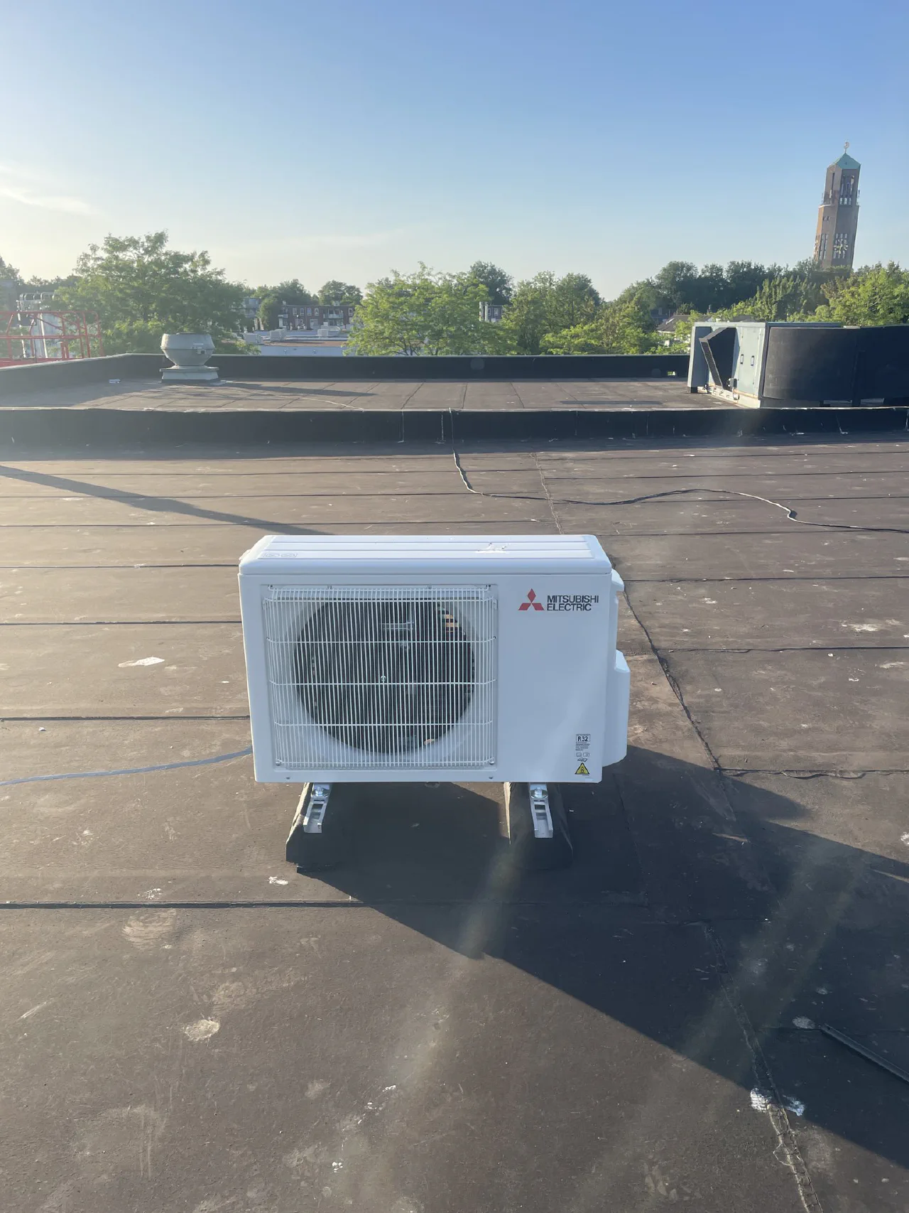 Mitsubishi Electric buitenunit op dakvoeten op een plat bitumen dak, met bomen en de Poldertoren van Emmeloord op de achtergrond