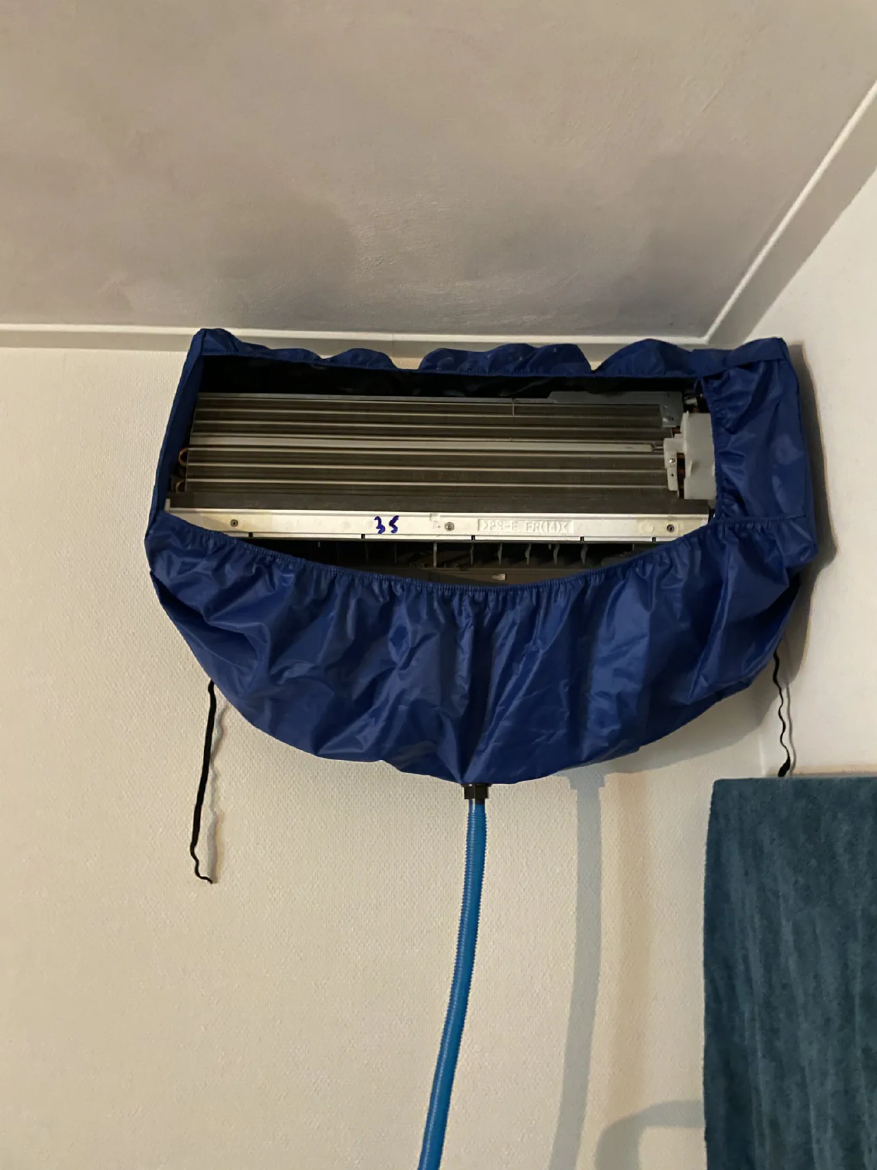 Open airco binnenunit aan de wand met een blauwe reinigingshoes eromheen en een blauwe afvoerslang naar beneden, verdamper en lamellen zichtbaar