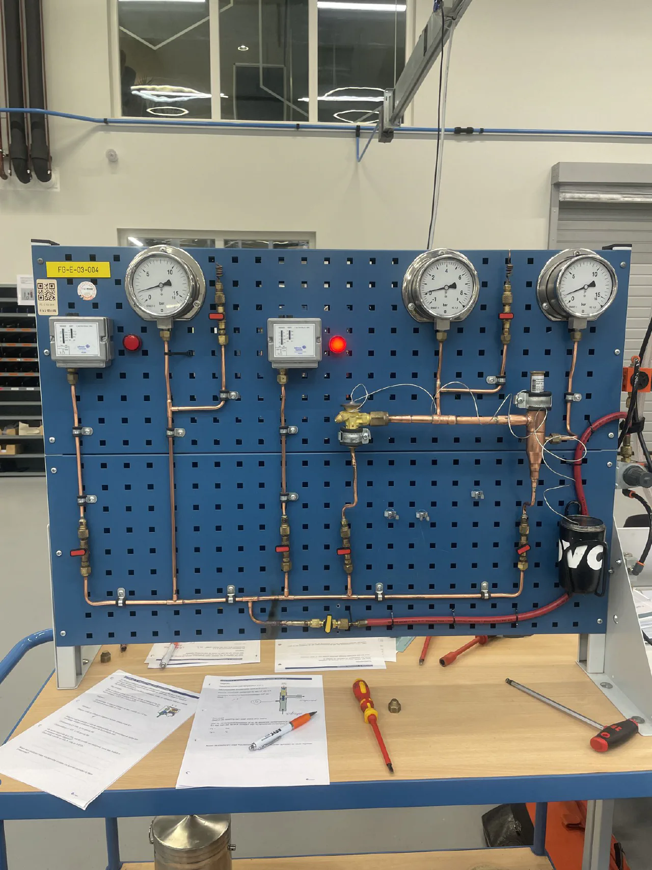 Blauw praktijkbord met vier manometers, twee Johnson Controls pressostaten met rode signaallampen, koperen leidingen, kogelkranen en een expansieventiel, werkbladen en schroevendraaiers op de tafel ervoor