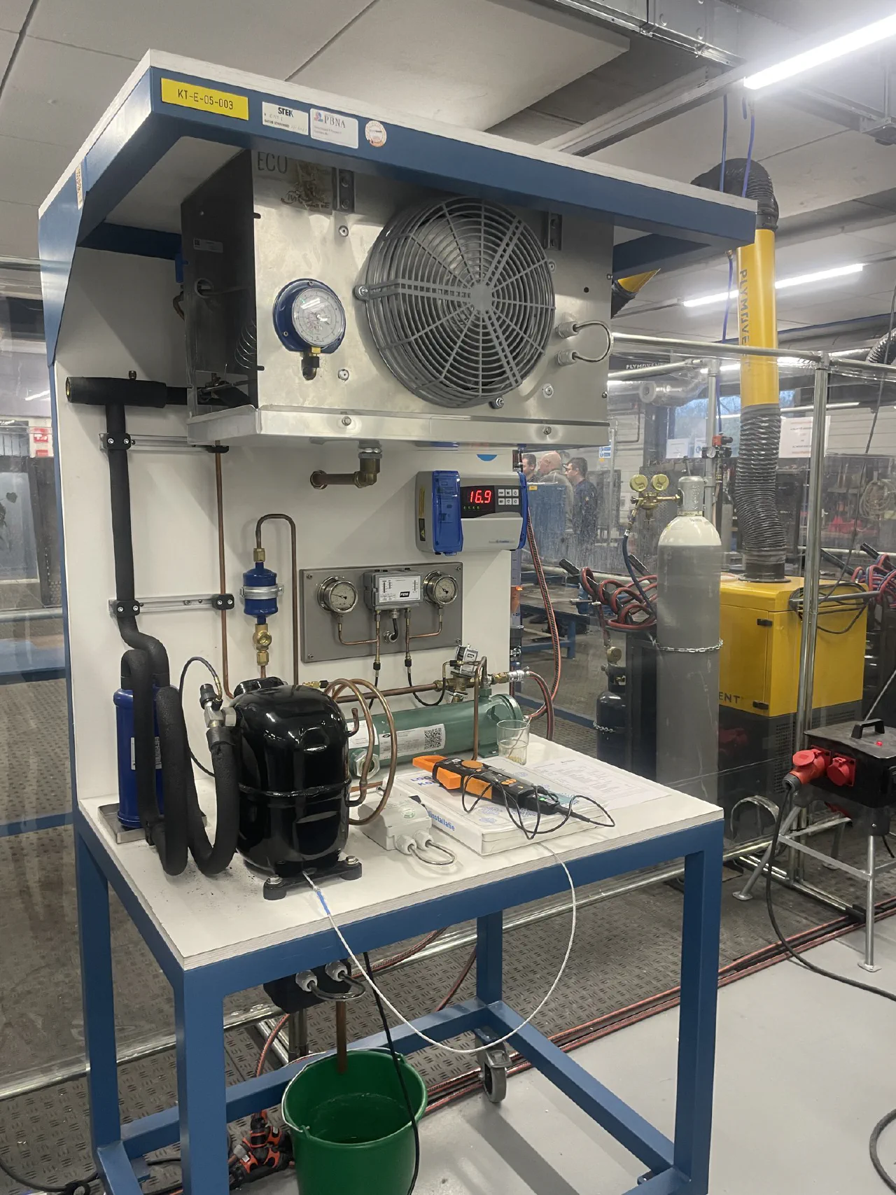 Complete oefenkoelinstallatie op een blauw frame met verdamper en ventilator bovenin, digitale regelaar met 16,9 graden, compressor, vloeistofvat, kijkglas en een stroomtang op het werkblad