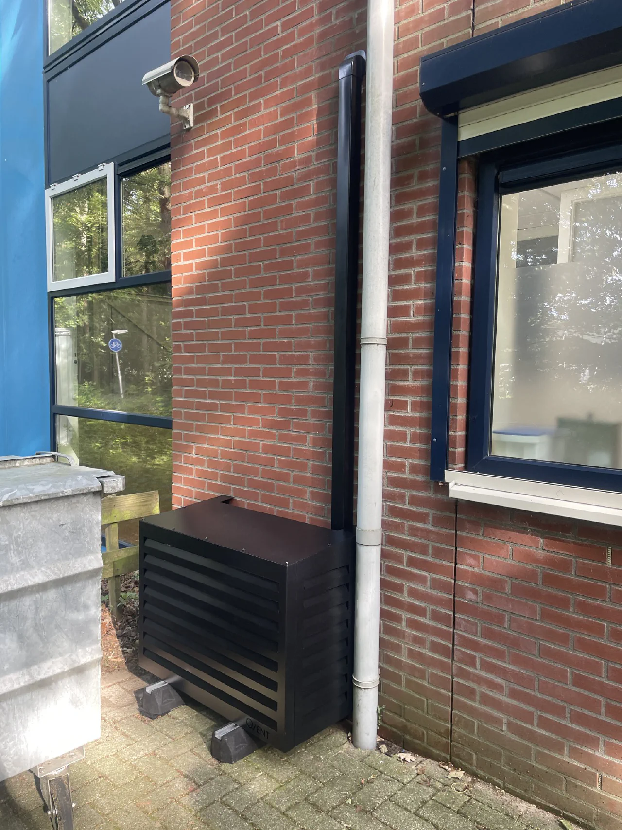 Zwarte Qvent omkasting met lamellen rond een buitenunit tegen een rode bakstenen gevel, zwarte leidinggoot omhoog naast een grijze regenpijp