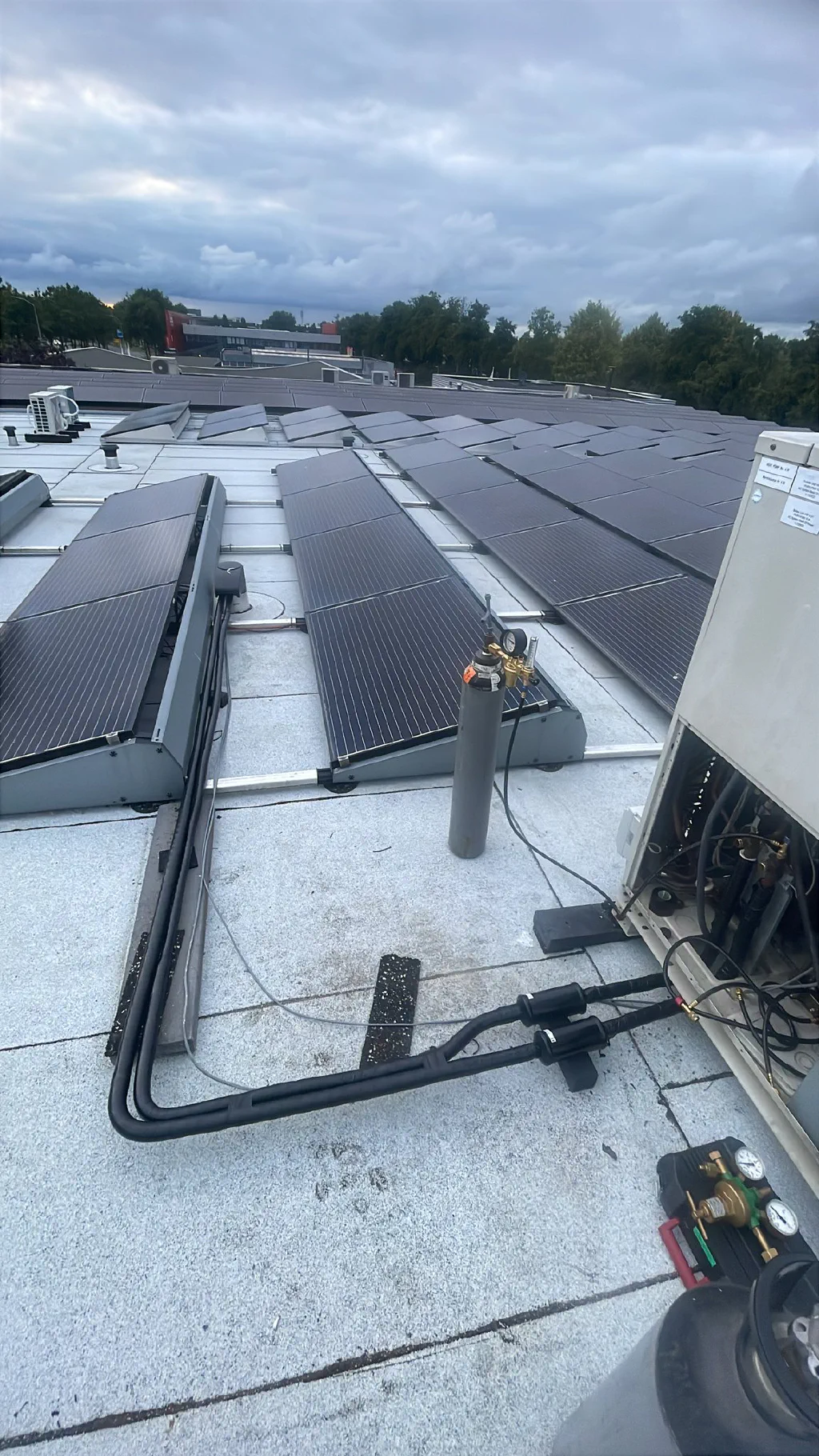 Plat dak met rijen zonnepanelen onder een bewolkte lucht, een grijze stikstofcilinder met reduceerventiel, zwart geïsoleerde koelleidingen en een geopende City Multi buitenunit met manometerset