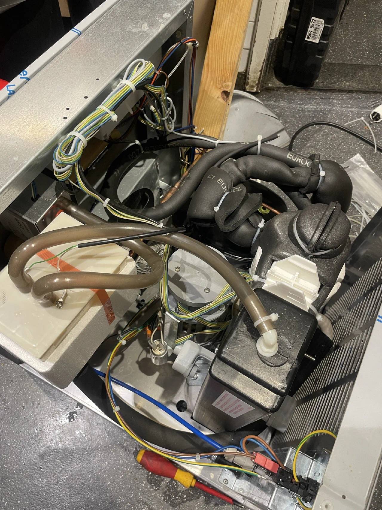 Geopend techniekcompartiment van een koelinstallatie met compressor, zwart geïsoleerde leidingen, bekabeling met kabelbinders en een schroevendraaier