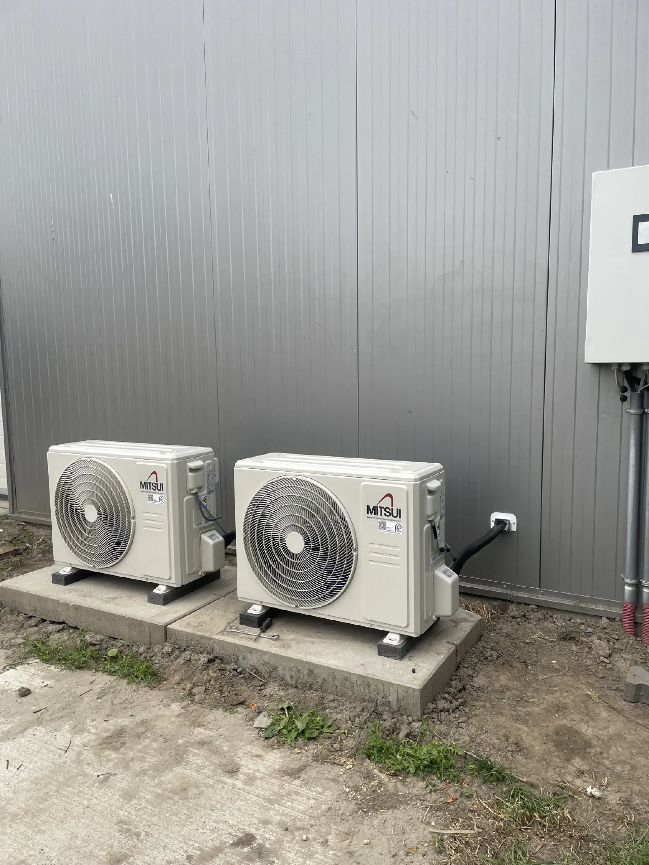 Twee witte Mitsui buitenunits op betontegels voor een grijze stalen gevel, met wanddoorvoer en buitenkast rechts